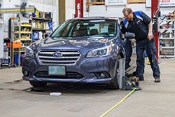 A mechanic adjusting the front wheel of a blue Subaru sedan in a shop.