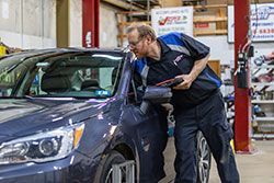 Mechanic examining a car's wheel alignment in a garage. He wears a work uniform and holds a tablet.
