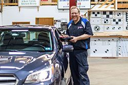 Man in mechanic uniform smiling next to a blue car inside a shop, holding a device.