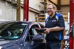 Mechanic in blue uniform smiles while holding a diagnostic tool next to a car in a repair shop.