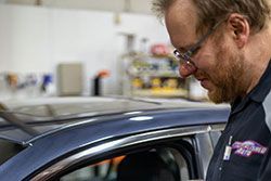 Mechanic in glasses inspecting a blue car's window in a garage.