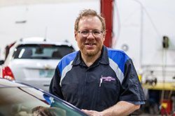 Mechanic in a blue and black uniform, smiles in auto repair shop; car and tools in background.