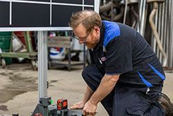 Man in work uniform kneeling, adjusting equipment near a black and white grid in a workshop.