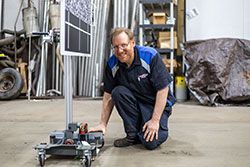 Man in blue uniform kneels beside a small robot with a screen, smiling in a workshop.