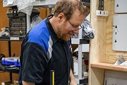Man in a blue and black work shirt working at a workbench, with equipment in the background.