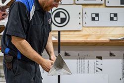 Person in a blue-accented mechanic's shirt working with a metal triangular object, likely for a test, with testing targets in the background.