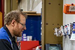 Mechanic looks at keys on a pegboard in a shop, wearing glasses and a blue-and-black work shirt.