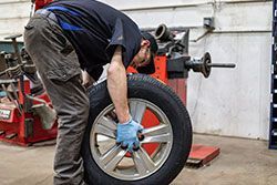 Mechanic in work clothes, inspecting a car tire at a repair shop, balancing machine in background.