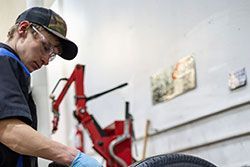 Mechanic in blue gloves works on tire in shop, wearing cap and safety glasses. Red tire machine visible.