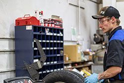 Mechanic in blue gloves balancing a tire at a workshop. He wears glasses and a cap, with shelves and tools in the background.