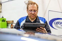 Mechanic in blue shirt using a tablet to diagnose a vehicle in a Cooper Tire shop.