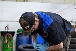 Mechanic wearing hat and gloves, leaning over tire in garage, inspecting.