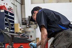 Mechanic working on equipment in a workshop; wearing a cap, blue gloves, and dark uniform.