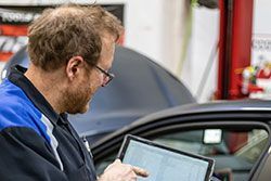 Mechanic in blue uniform uses a tablet to diagnose a car, inside a shop.