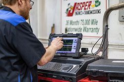 Mechanic using a diagnostic tool to examine a car. Tool displays graphs, with Jasper warranty sign in the background.