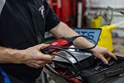 Mechanic holding a red diagnostic tool with a black cable, near a laptop and toolbox.