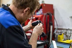 Mechanic in blue and black shirt using a tool with a computer in background.
