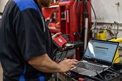 Mechanic uses diagnostic tool connected to laptop, likely to troubleshoot a vehicle's computer system in a workshop.