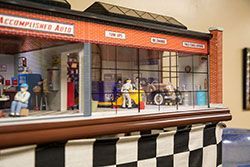 Miniature model of a classic auto repair shop interior, with small figures and a car, visible through large windows.