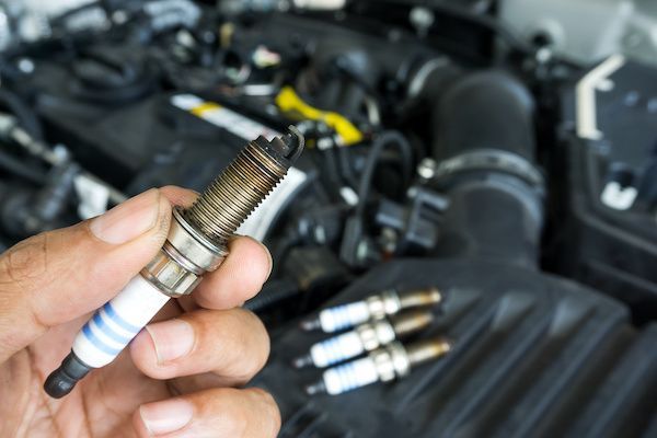 Hand holding a used spark plug, car engine bay in background, other plugs visible.