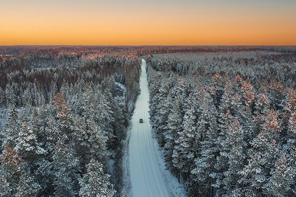 Snow-covered road through a forest at sunset, car driving. Orange and blue sky.