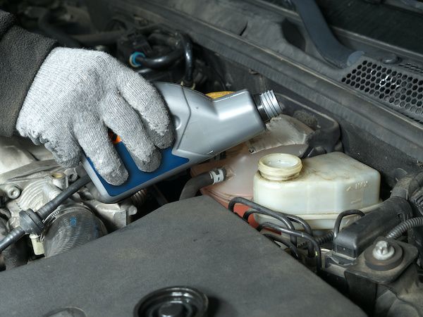 A gloved hand pouring brake fluid into a car's reservoir under the hood.