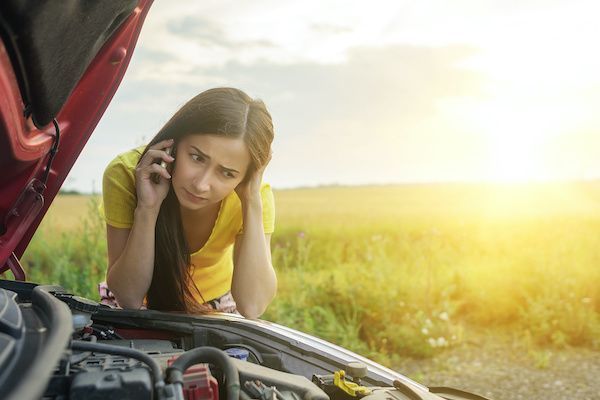 Woman with car trouble on the roadside, looking at engine, on phone, with worried expression.