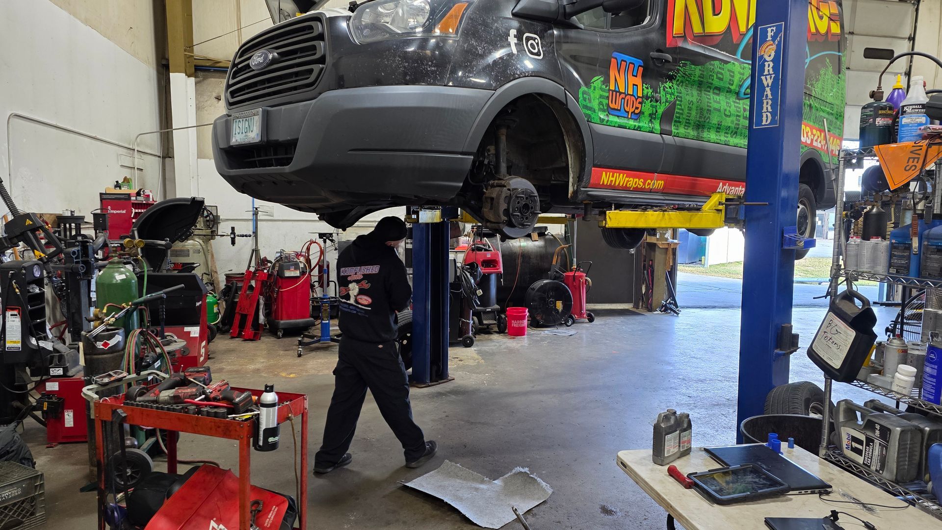 Mechanic working under a van raised on a lift in a repair shop. Tools and equipment surround him. | Accomplished Automotive