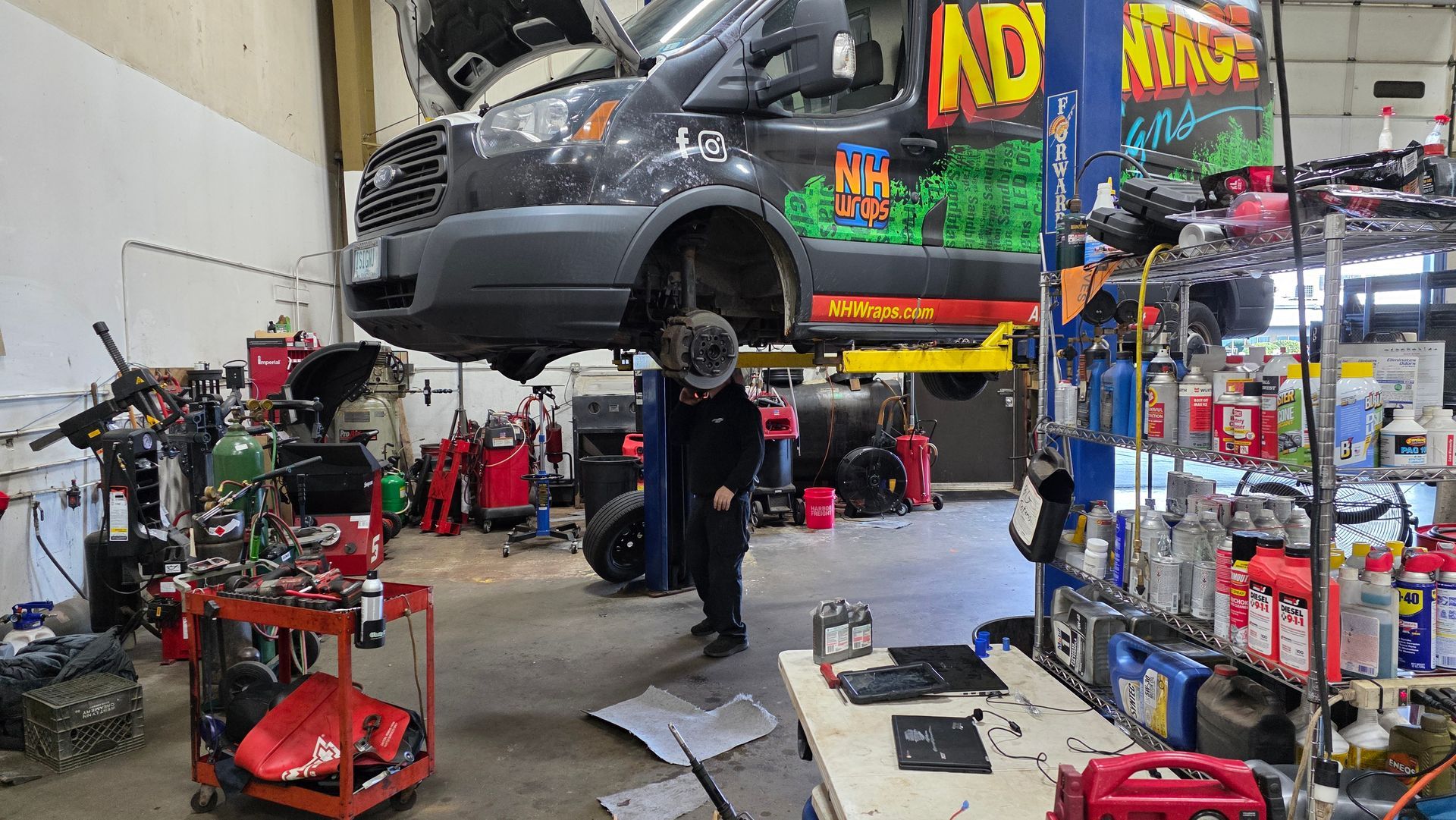Mechanic working under a lifted van in a cluttered auto repair shop. The van has business graphics. | Accomplished Automotive
