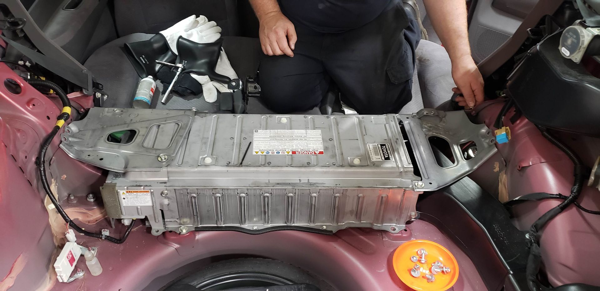 A hybrid car battery being worked on by a person. The battery is silver and rectangular. The car's interior is visible. | Accomplished Automotive