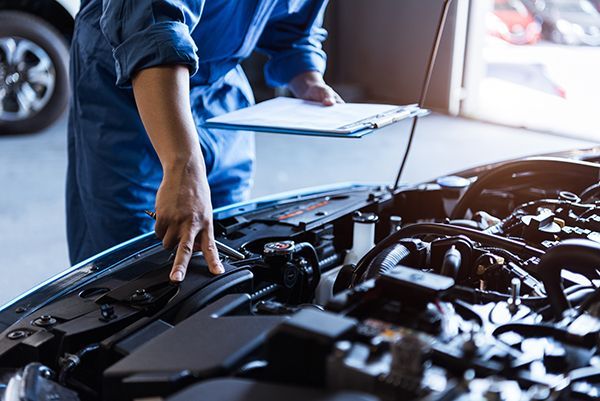 Mechanic in blue jumpsuit examining car engine, holding clipboard in a garage.
