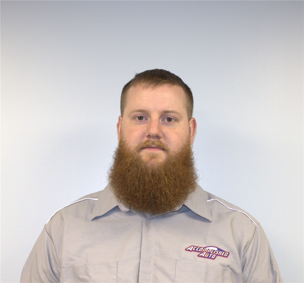 Man with a full beard wearing a grey uniform shirt, looking forward in front of a white wall.