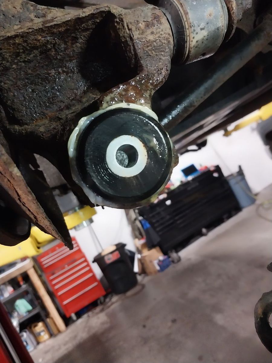 Close-up of a black and white rubber bushing installed on a rusty car suspension arm. Garage setting. | Accomplished Automotive