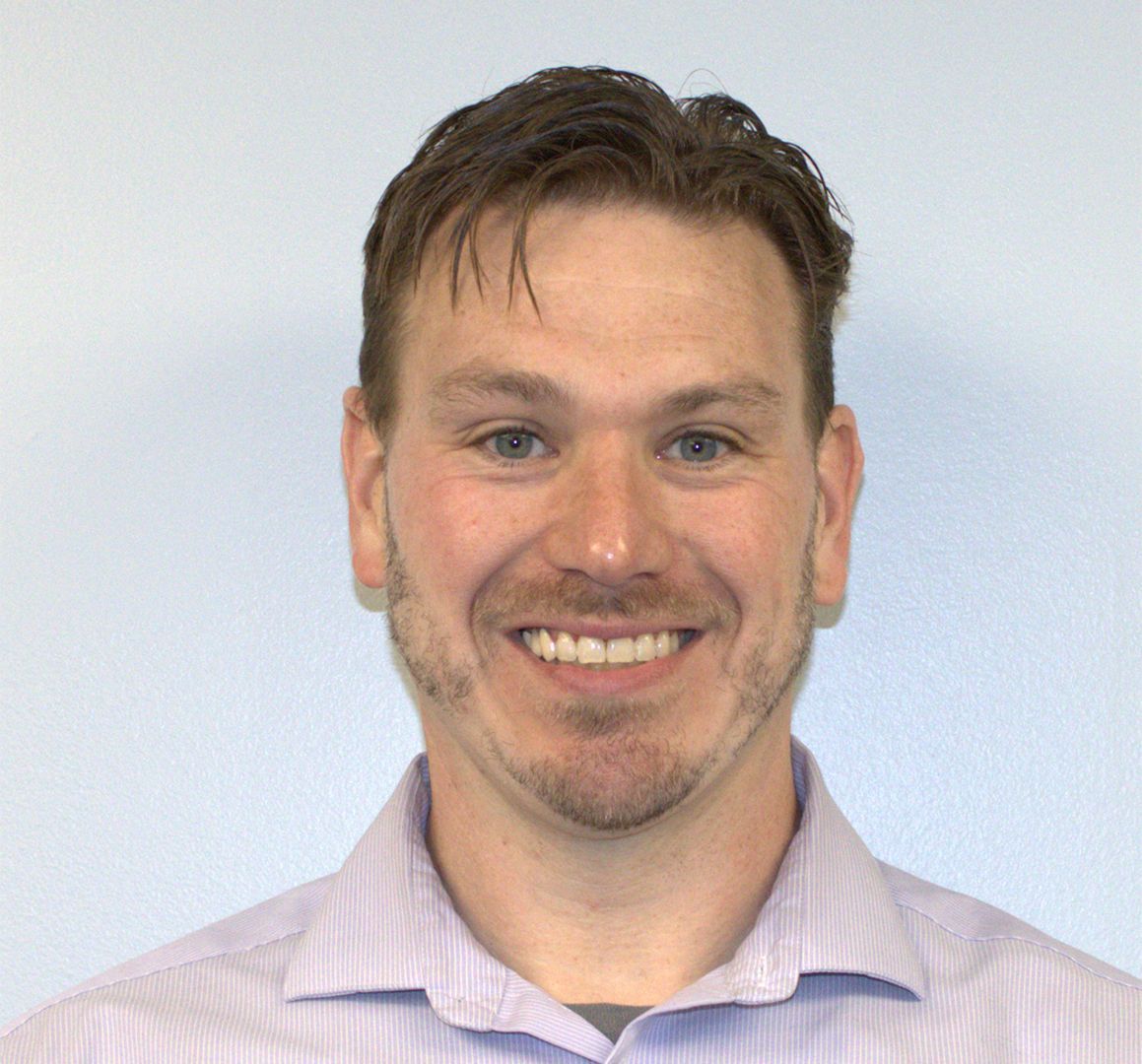 Man smiling at the camera, wearing a light purple button-down shirt, neutral background.