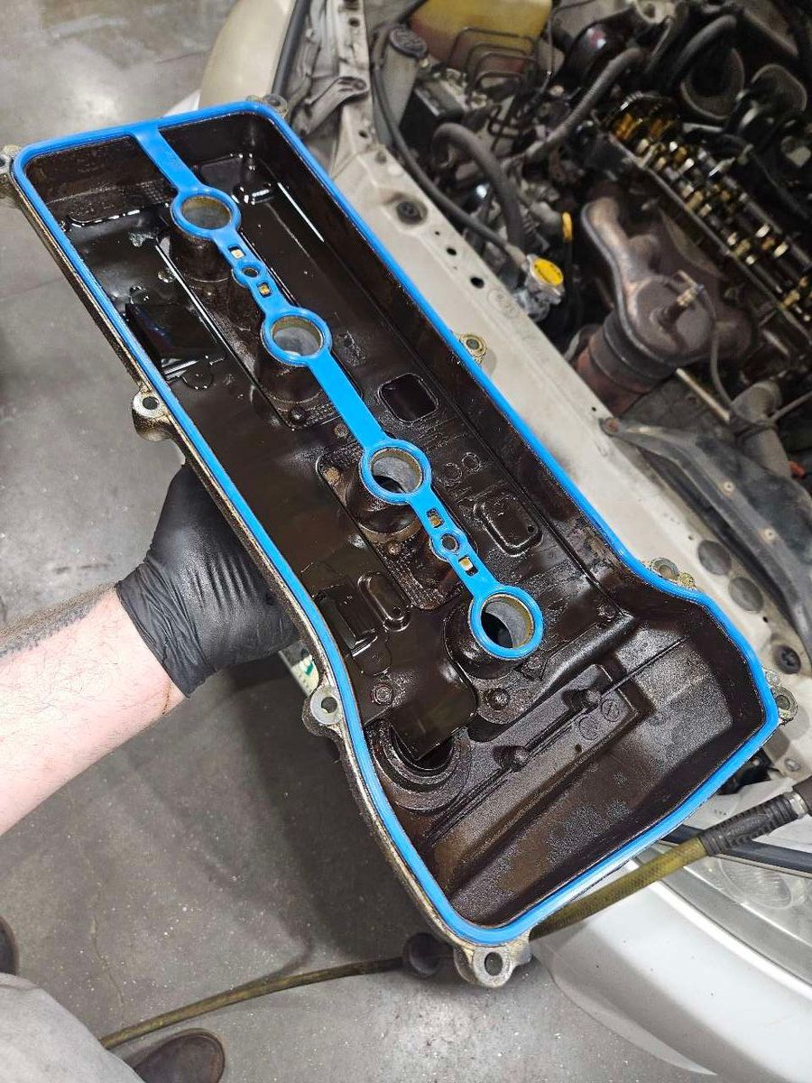 Hand holding a black and oily engine valve cover with a blue gasket, next to an engine. | Accomplished Automotive