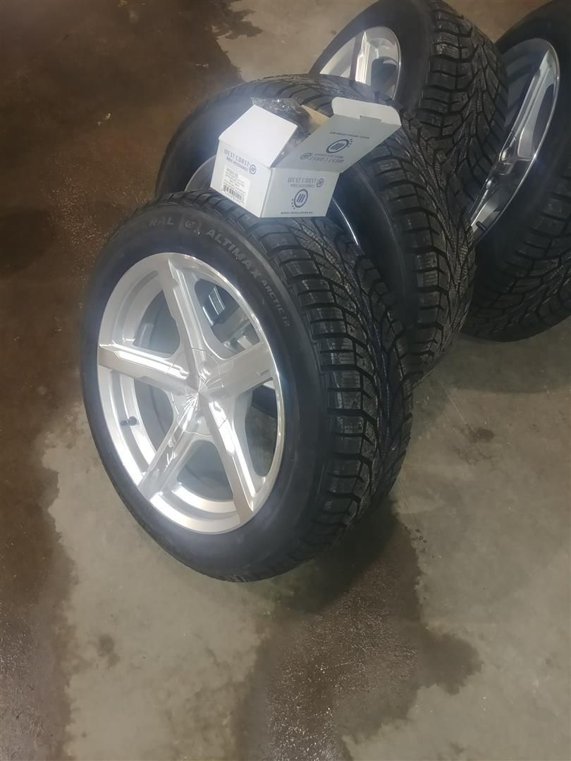 Four car wheels with silver rims and tires, stacked on a gray floor.