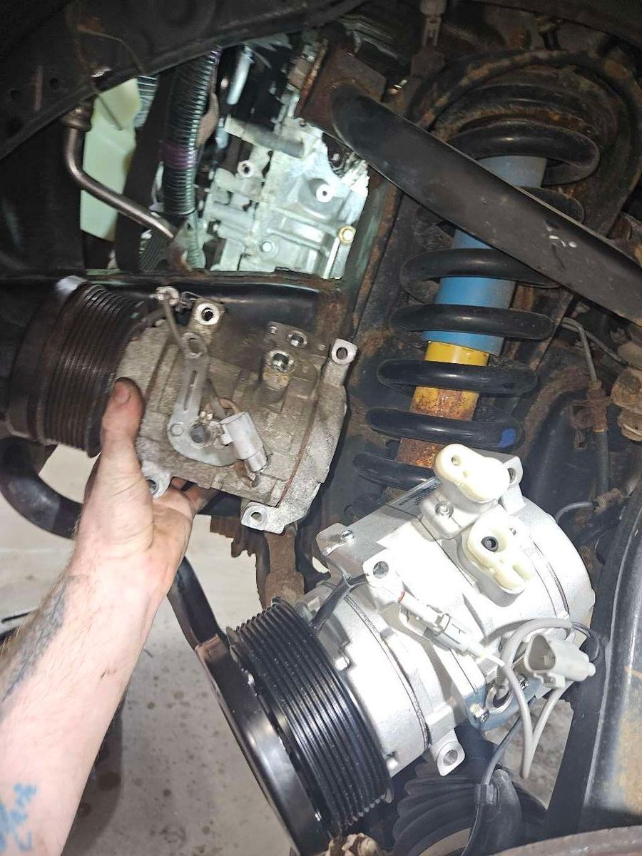 Person's hand holding an AC compressor next to another, mounted in a vehicle engine bay. | Accomplished Automotive