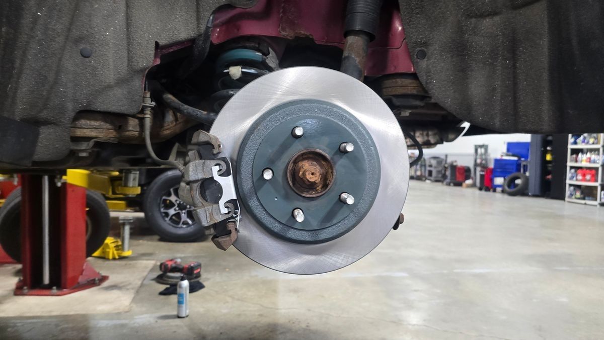 Car's rear brake disc is exposed, mounted with a caliper. It's in a garage, likely being serviced. | Accomplished Automotive