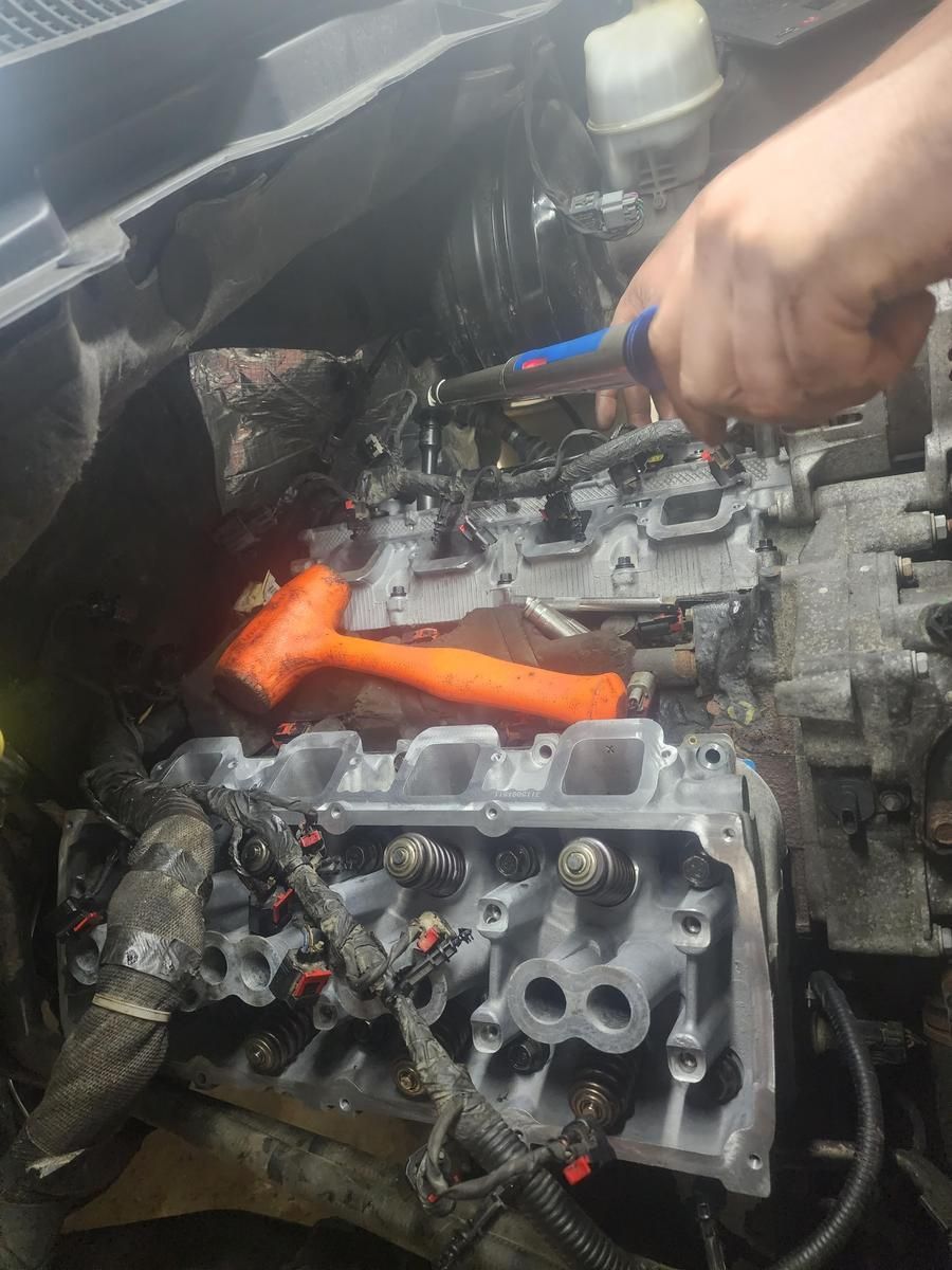 Mechanic working on an engine with a hammer and wrench. The engine is gray and exposed in an open car bay. | Accomplished Automotive