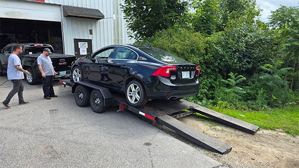 Black car on a trailer being loaded by two people outside a garage.