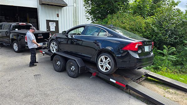 Black Volvo sedan on a trailer being loaded by a person in front of a building with a truck nearby.
