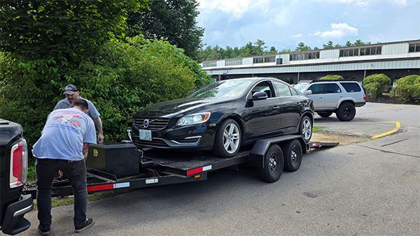 Two men loading a black Volvo onto a trailer, parked near a building and a green bush.