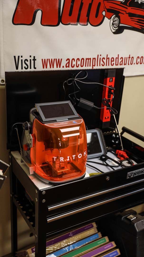 An orange car diagnostic tool sits on a black tool cart in an auto shop with a website banner.