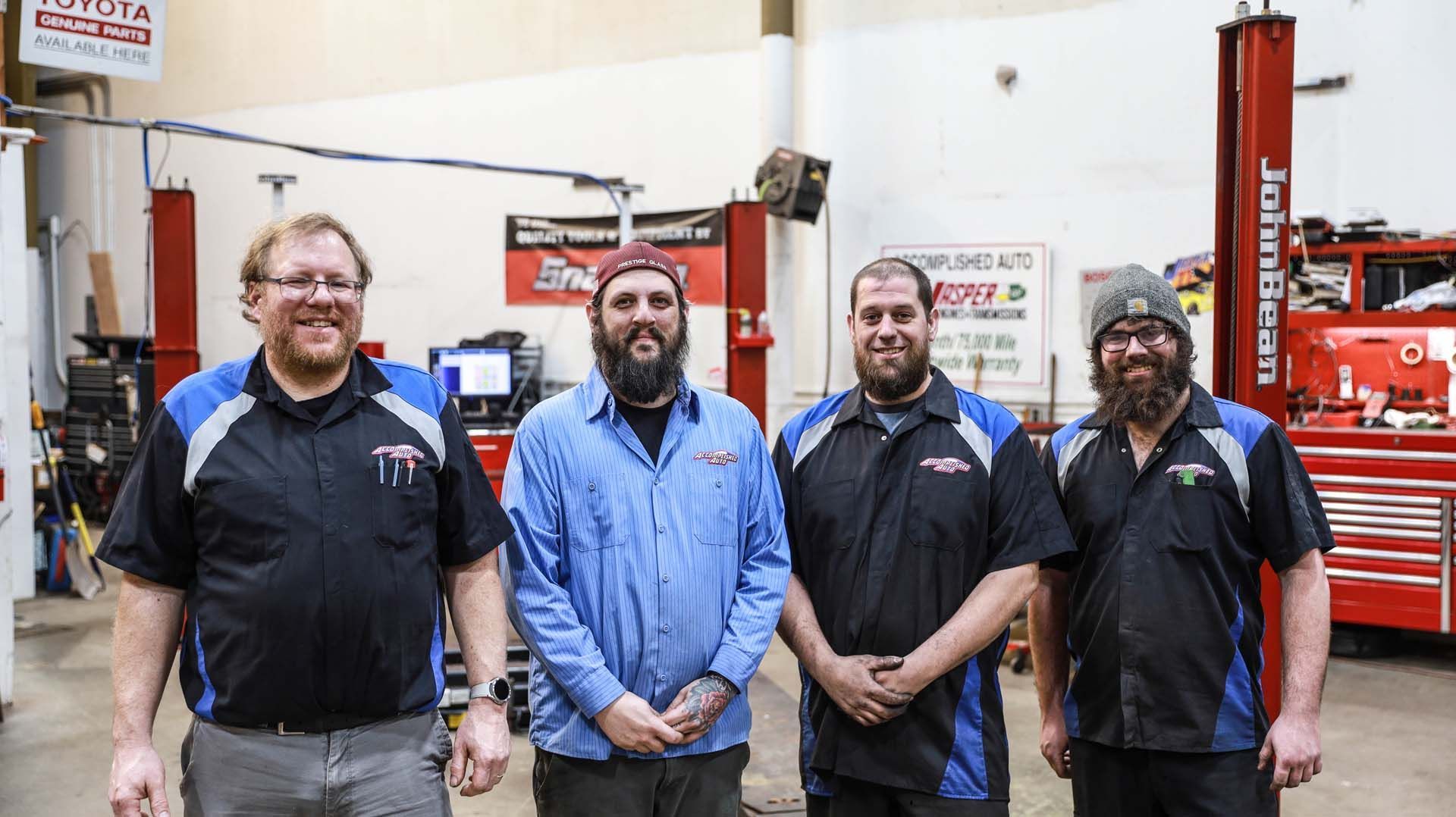 Four mechanics in a garage stand, smiling. They wear shop uniforms. Behind them are tools and equipment.