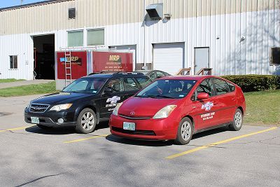 Two cars parked in front of a light-colored building. One is red, the other black. Both have logos on the doors.