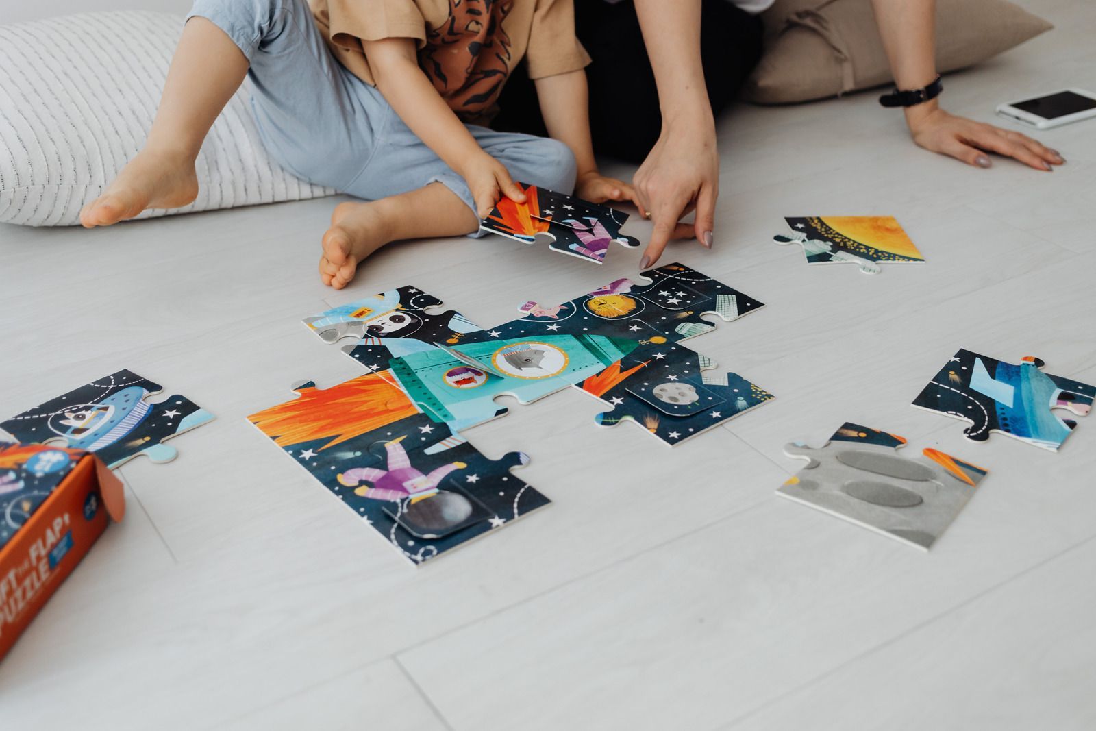 Child and adult assembling a space-themed puzzle on a light floor, several puzzle pieces scattered.