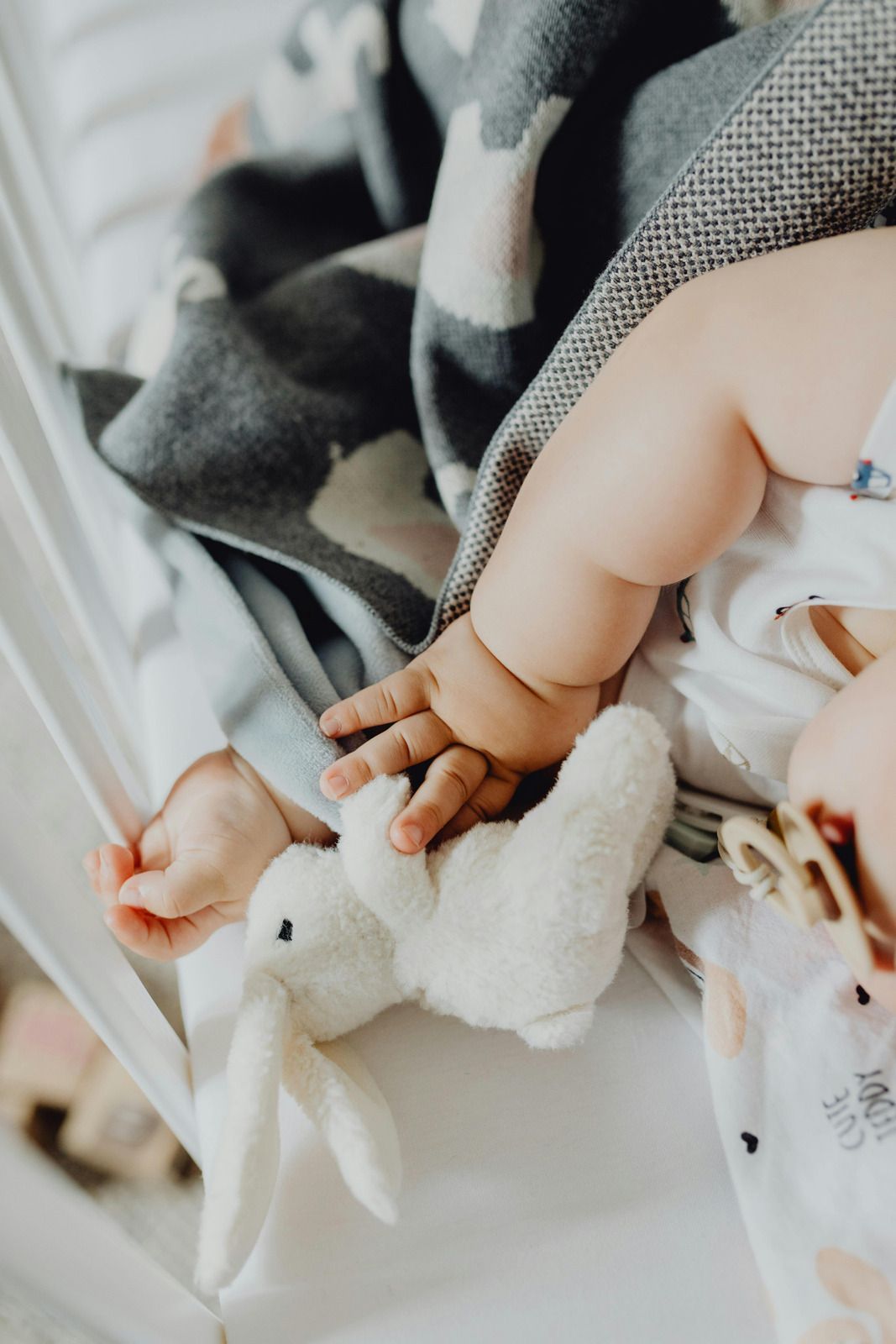 Baby's arm reaching for a white stuffed bunny in a crib, covered by a gray blanket.