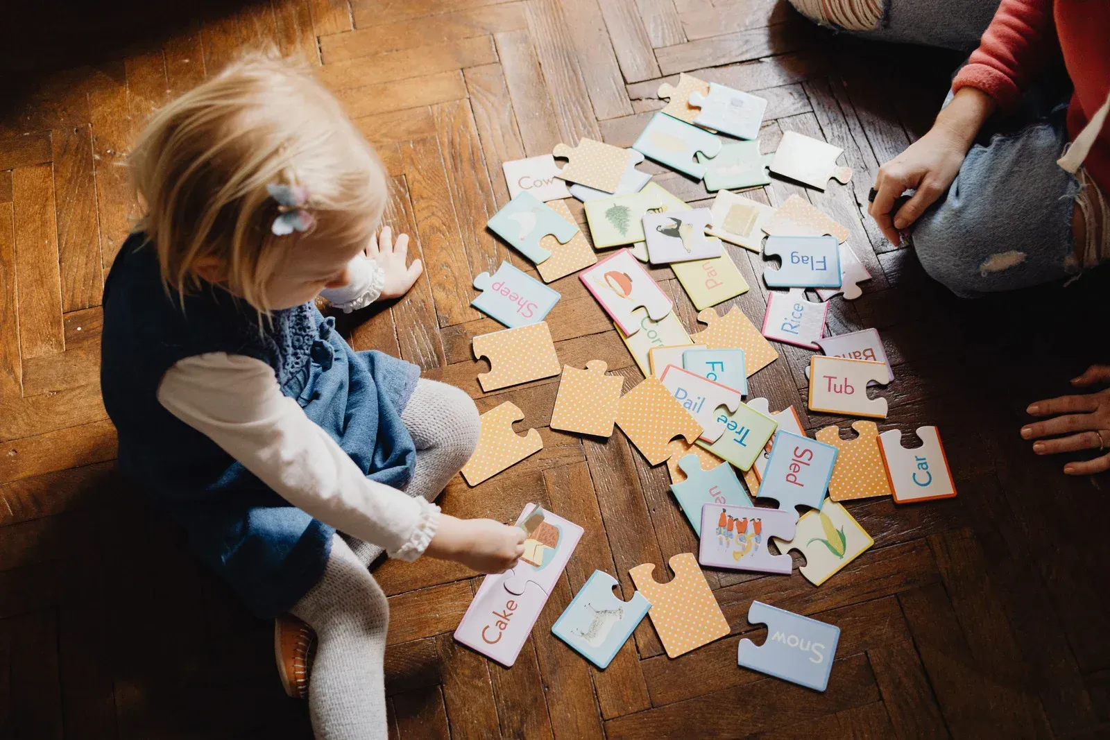 Child assembling colorful puzzle pieces with letters on a wooden floor, assisted by an adult.