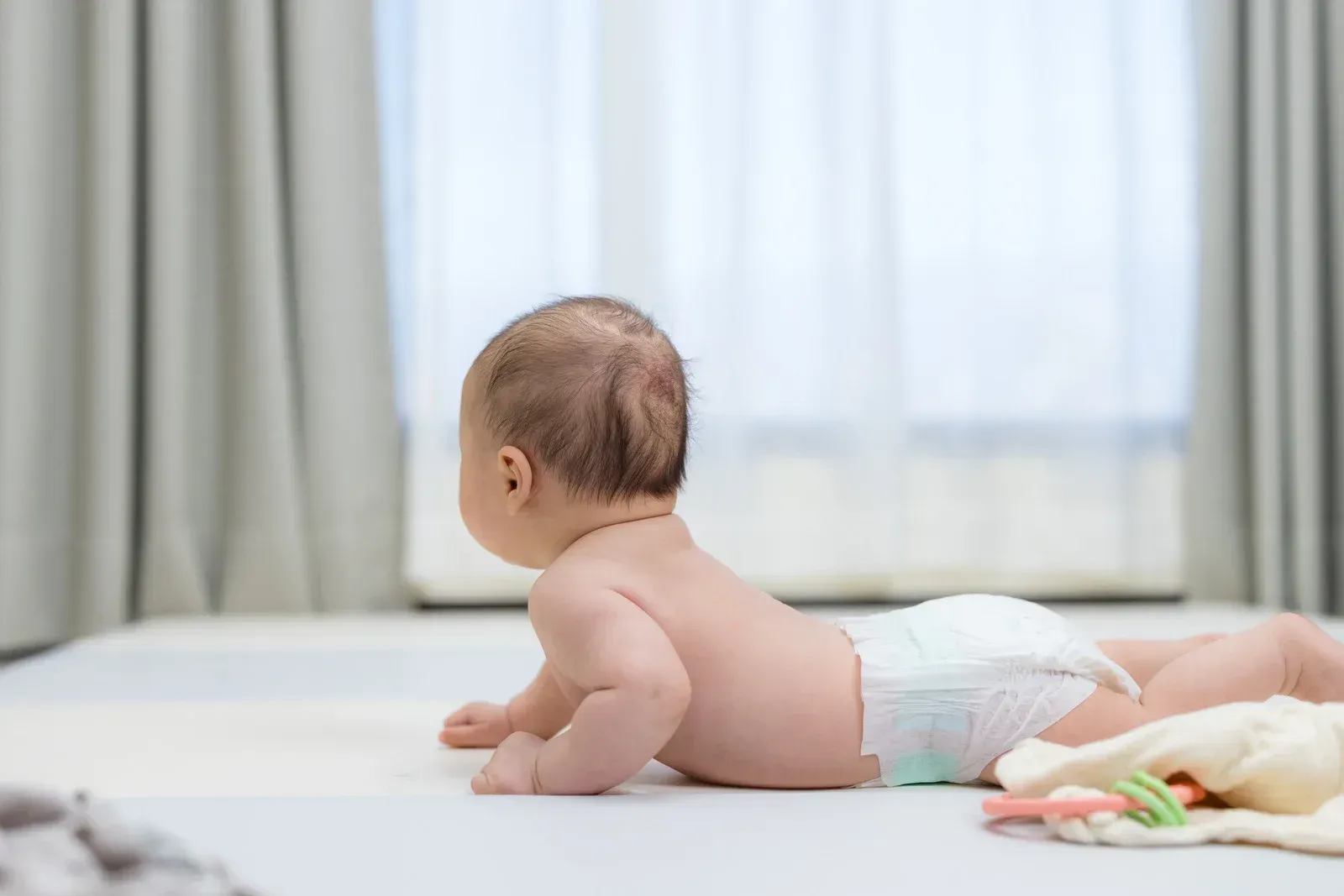 Baby on stomach wearing diaper on bed, looking toward a window.