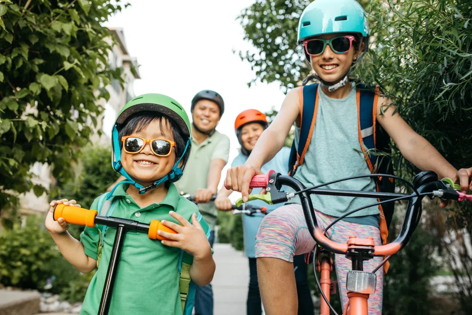 Children wearing helmets and sunglasses on bikes and scooters outdoors.